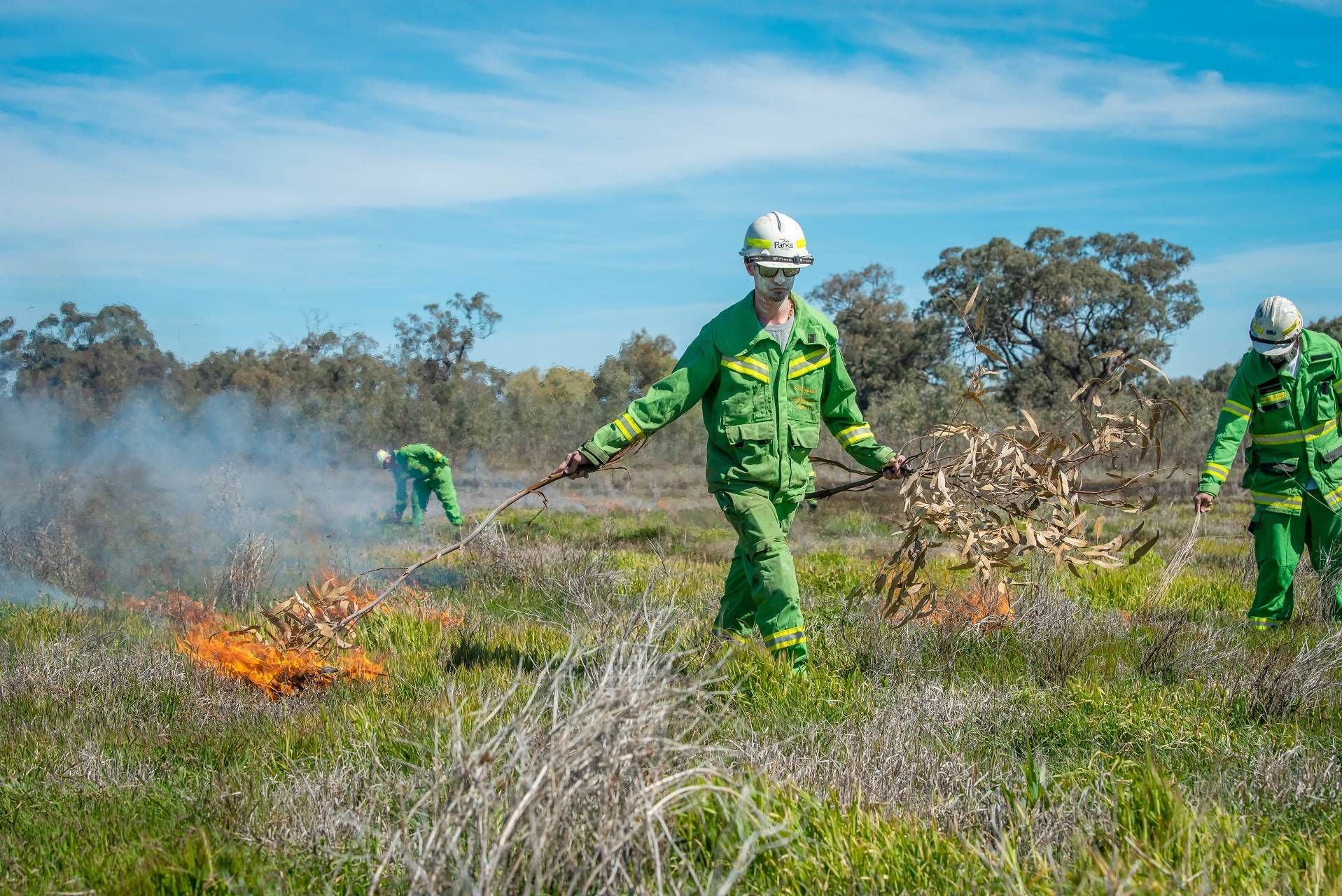 Man dressed in green overalls and wearing hard hat is igniting grasses using a branch that is on fire.