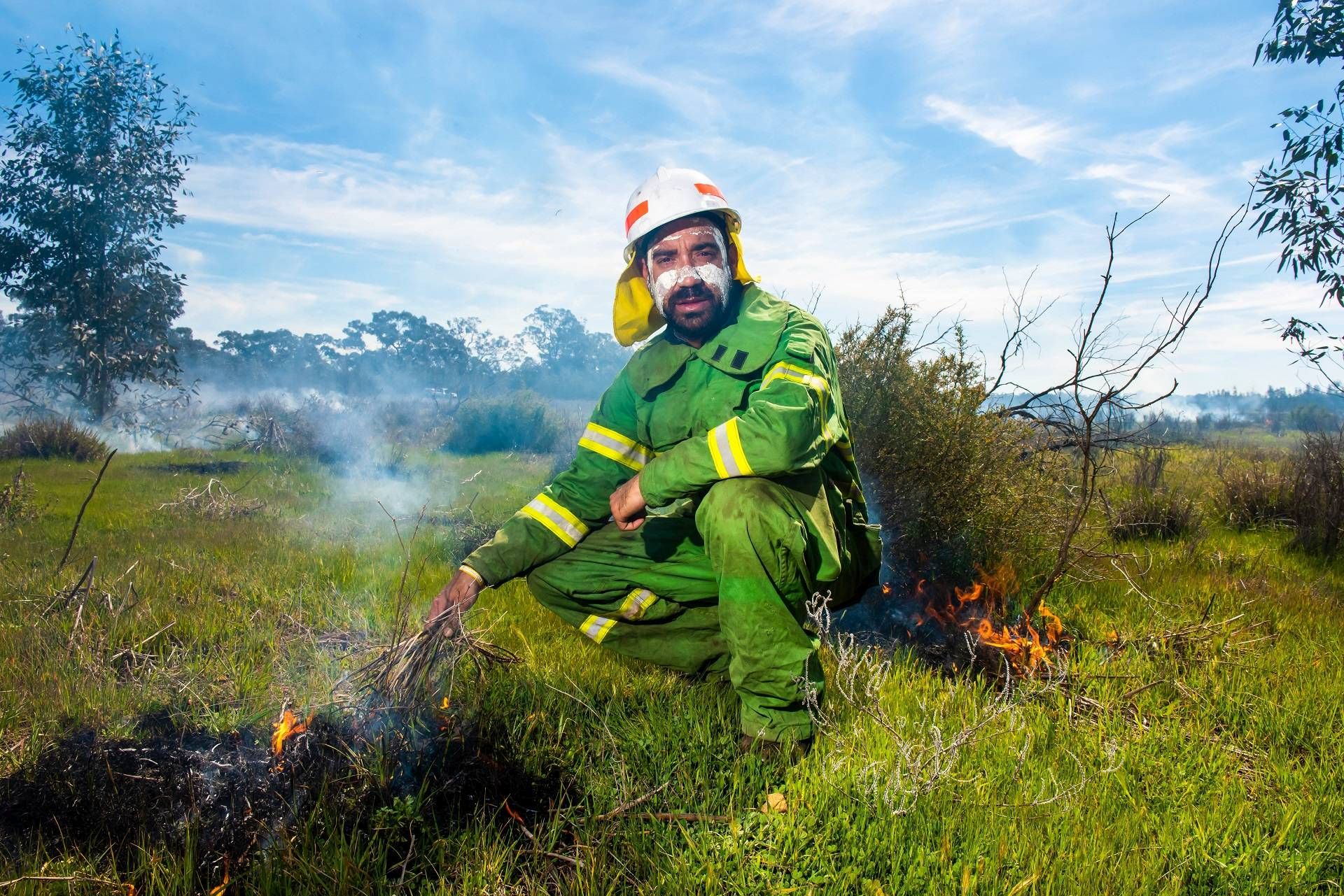 Man wearing a hard hat and green overalls is crouching and looking directly at the camera. In his right hand he is holding dried grasses which he is using to ignite a Cultural Burn. Behind him is smoke and flames.