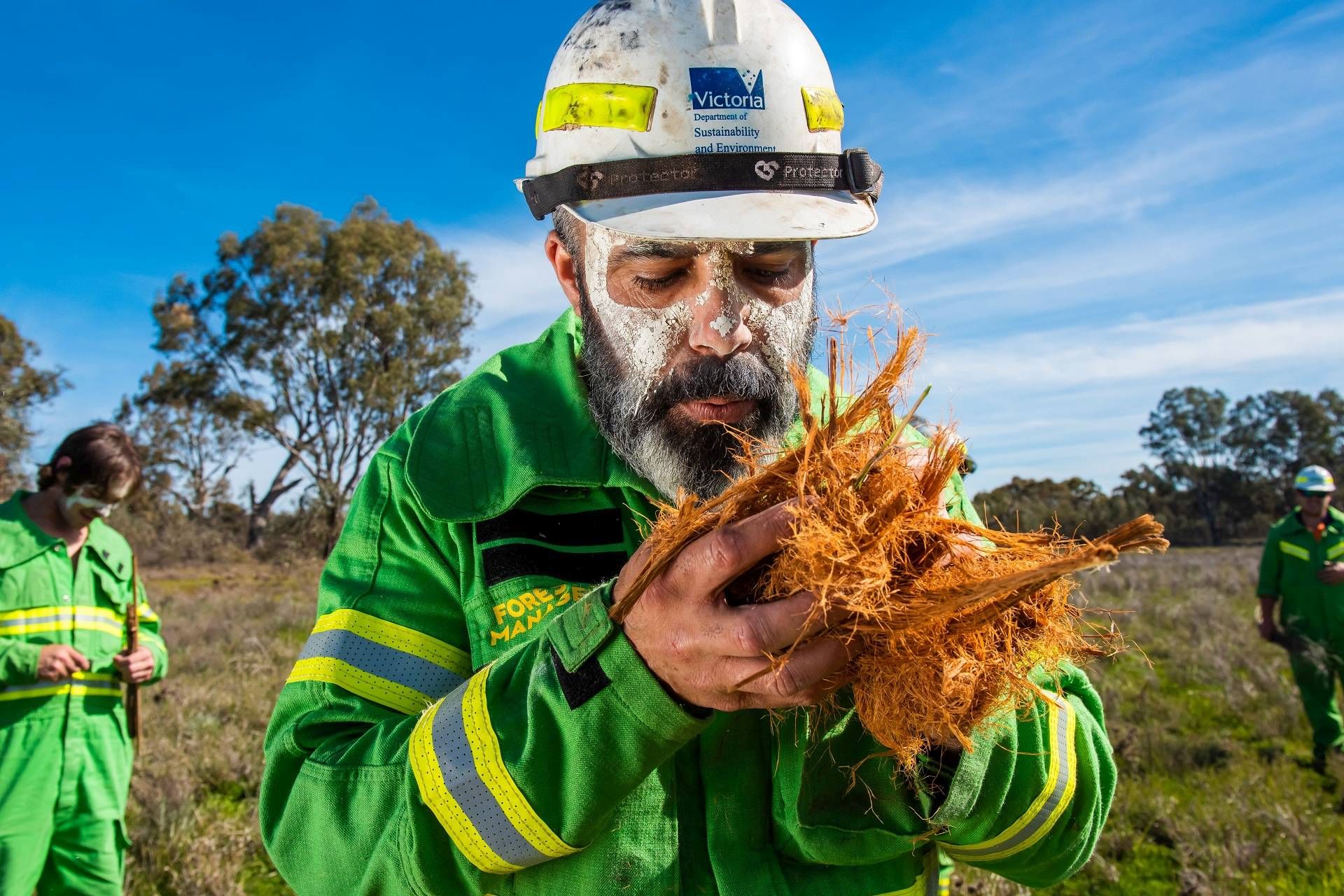 Man wearing a hard hat and green overalls is blowing on brown husks to try and create fire.